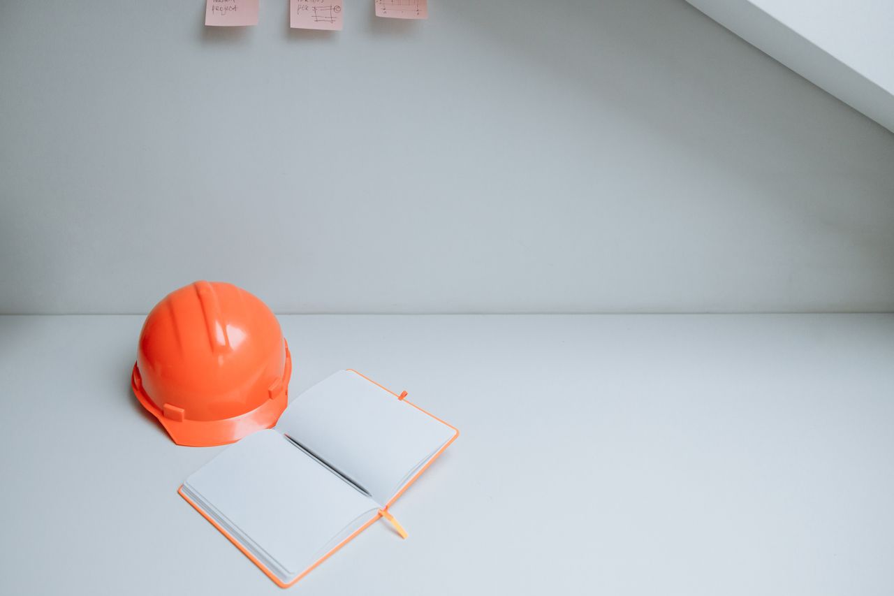 Minimalist workspace with orange hardhat and open notebook on a white desk, perfect for concept design.